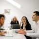 A group of four colleagues in a tense meeting, with two employees in the foreground actively arguing, while a third listens and a fourth appears stressed in the background.