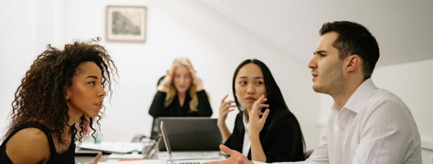 A group of four colleagues in a tense meeting, with two employees in the foreground actively arguing, while a third listens and a fourth appears stressed in the background.