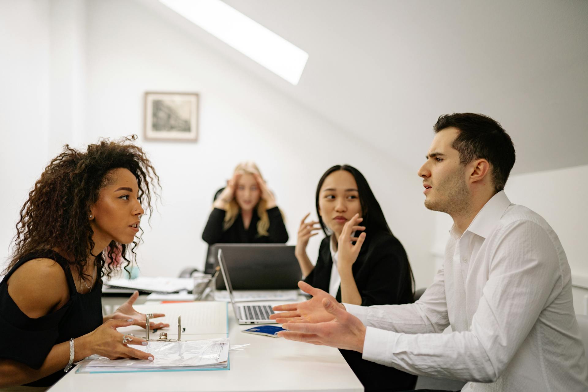 A group of four colleagues in a tense meeting, with two employees in the foreground actively arguing, while a third listens and a fourth appears stressed in the background.